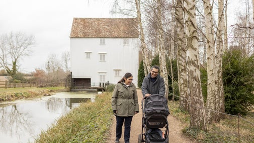 A couple pushing a pram walk beside a stream with a white wooden slatted building behind them in winter